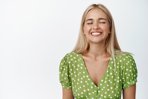 Bezpłatne zdjęcie beztroska blond dziewczyna zamyka oczy, uśmiecha się i czuje się szczęśliwy, oddychając, stojąc w zielonej sukience na białym tle