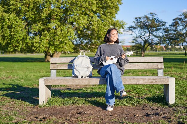 Beztroska dziewczyna siedzi na ławce w parku z ukulele gra i śpiewa na świeżym powietrzu w słoneczny szczęśliwy dzień