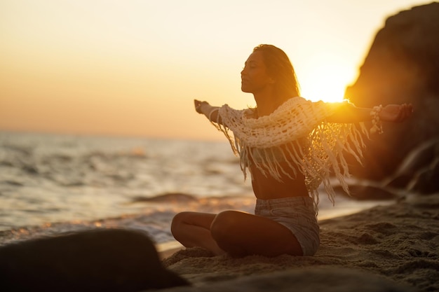 Beztroska kobieta z wyciągniętymi ramionami, ciesząca się wolnością podczas relaksu na piasku na plaży podczas zachodu słońca