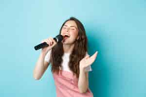 Bezpłatne zdjęcie beztroska piękna kobieta wykonuje piosenkę, z pasją śpiewa do mikrofonu, gra karaoke, stoi na niebieskim tle