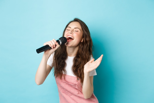 Beztroska piękna kobieta wykonuje piosenkę, z pasją śpiewa do mikrofonu, gra karaoke, stoi na niebieskim tle