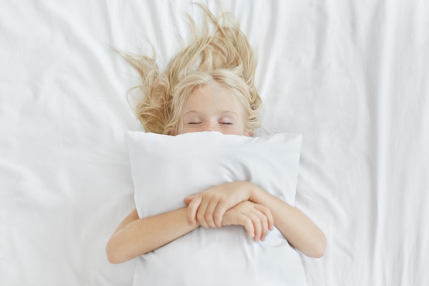 Beztroska, spokojna dziewczynka leżąca na białej pościeli, obejmująca poduszkę, mając jednocześnie przyjemne sny. Blondynka z piegami śpi w łóżku po spędzeniu całego dnia na pikniku. Spokojne dziecko