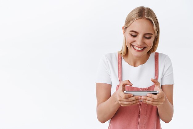 Beztroska szczęśliwa młoda kaukaska blond dziewczyna w kombinezonie tshirt trzymaj smartfon poziomo i śmiejąc się, grając śmieszną aplikację pobierz nową fajną grę dotknij ekranu przyciski stoją białe tło