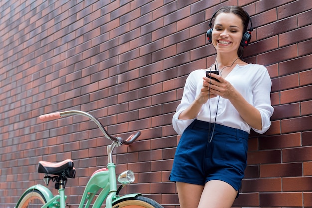 Beztroski czas. piękna młoda kobieta w słuchawkach, słuchająca odtwarzacza mp3 i uśmiechająca się, stojąc w pobliżu swojego zabytkowego roweru