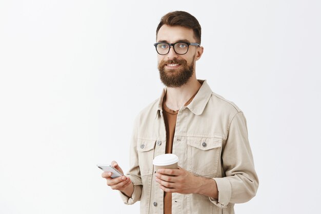 Beztroski przystojny brodaty mężczyzna w okularach pozuje przy białej ścianie
