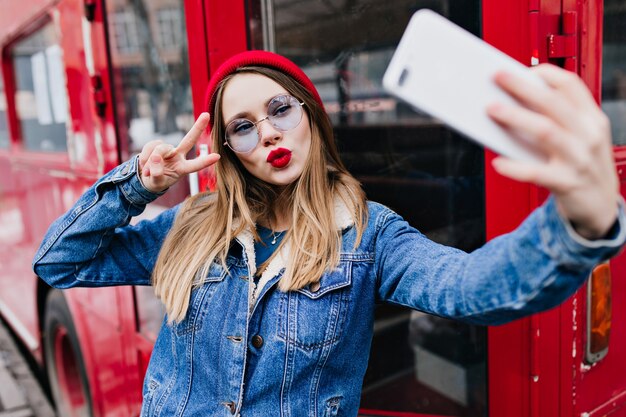 Biała dziewczynka kaukaski przy użyciu telefonu do selfie podczas całowania wyrazu twarzy. Pani na zewnątrz w dżinsowej kurtce i czerwonym kapeluszu stojąca na ulicy i robiąca sobie zdjęcie.