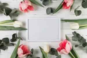 Bezpłatne zdjęcie biała pusta rama otaczająca z białymi i różowymi tulipanami na drewnianym biurku