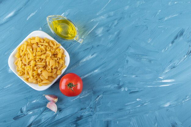 Biała tablica surowego makaronu z olejem i świeżymi czerwonymi pomidorami na niebieskim tle.