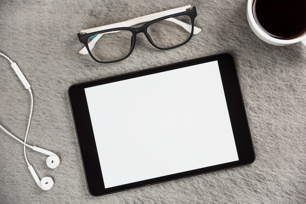 Białe słuchawki; okulary; filiżanka kawy i pusty ekran cyfrowy tablet na szarym biurku