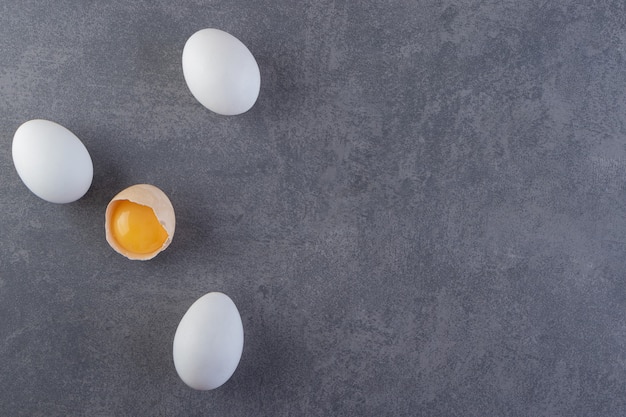 Białe surowe jajka i popękane jajko umieszczone na kamiennym stole.