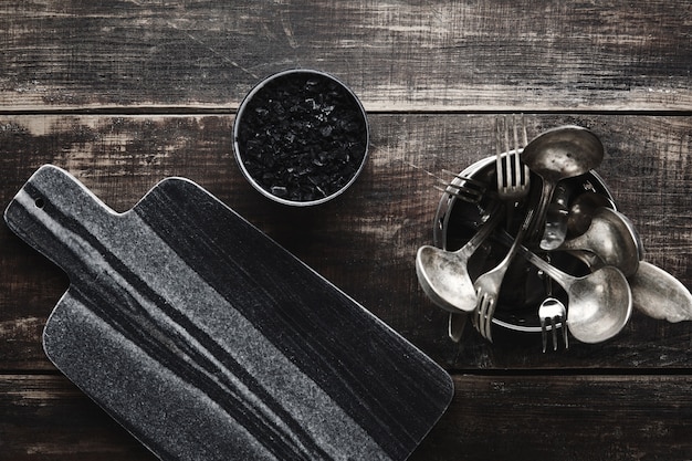 Bezpłatne zdjęcie biurko z czarnego marmuru, sól wulkaniczna i zabytkowe wyroby kuchenne: widelec, nóż, łyżka w stalowym garnku na postarzanym drewnianym stole. widok z góry.