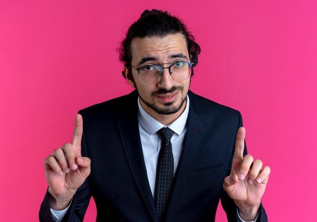 Biznes człowiek w czarnym garniturze i okularach patrząc do przodu pokazując palce wskazujące stojąc na różowej ścianie
