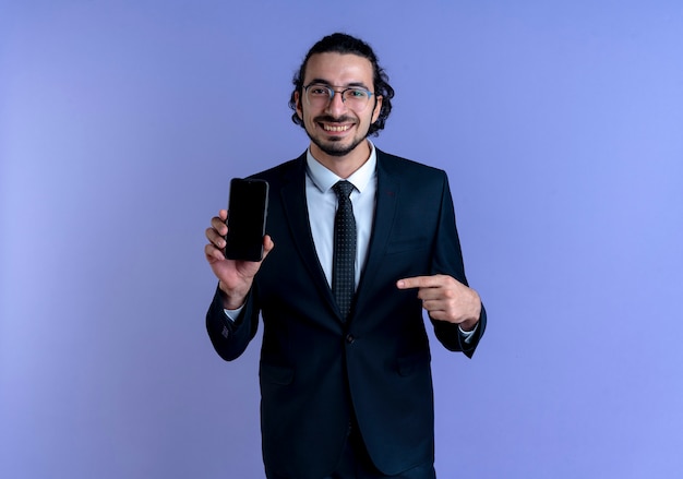 Biznes człowiek w czarnym garniturze i okularach pokazuje smartfon, wskazując palcem na to uśmiechnięty pewnie stojący nad niebieską ścianą