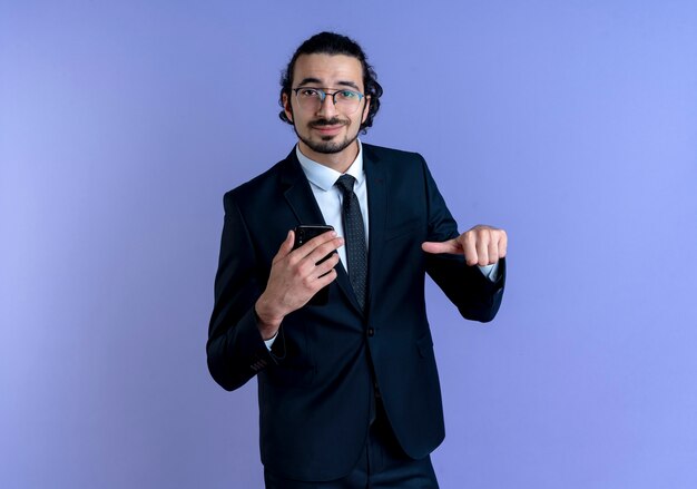 Biznes człowiek w czarnym garniturze i okularach pokazuje smartfon, wskazując palcem na to uśmiechnięty pewnie stojący nad niebieską ścianą