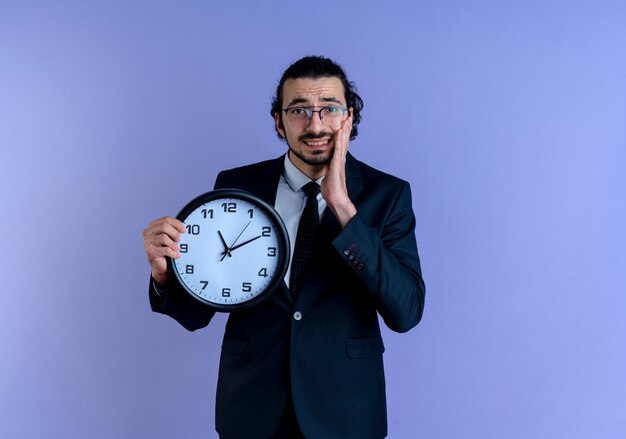 Biznes człowiek w czarnym garniturze i okularach trzyma zegar ścienny patrząc zdezorientowany stojąc nad niebieską ścianą