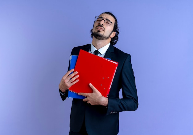 Biznes człowiek w czarnym garniturze i okularach, trzymając foldery z zamkniętymi oczami, zmęczony i znudzony stojąc nad niebieską ścianą