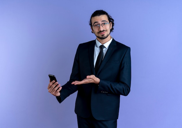 Biznes człowiek w czarnym garniturze i okularach, trzymając smartfon, prezentując z ramieniem ręki uśmiechnięty pewnie stojący nad niebieską ścianą