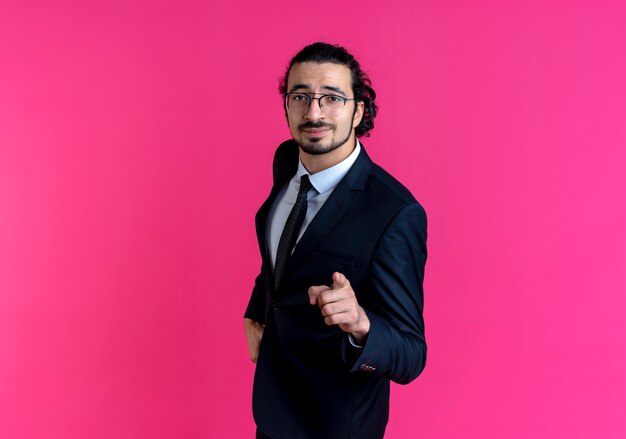 Biznes człowiek w czarnym garniturze i okularach, wskazując palcem do przodu, patrząc pewnie stojąc na różowej ścianie