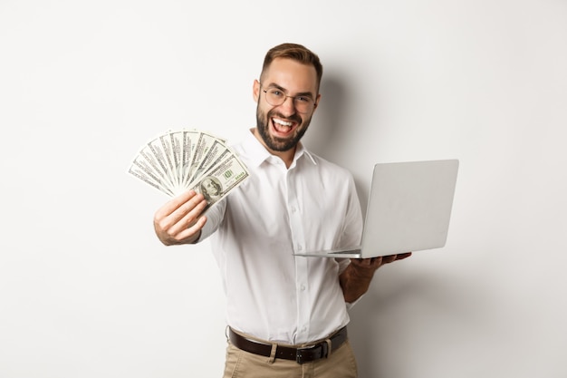 Biznes i e-commerce. Szczęśliwy odnoszący sukcesy biznesmen chwalący się pieniędzmi, pracujący na laptopie online, na stojąco