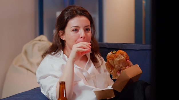 Biznes kobieta oglądając telewizję jedzenie hamburgera w salonie po pracy. młody dorosły cieszący się posiłkiem typu fast food na obiad, siedząc na kanapie i śmiejąc się z ekranu telewizora