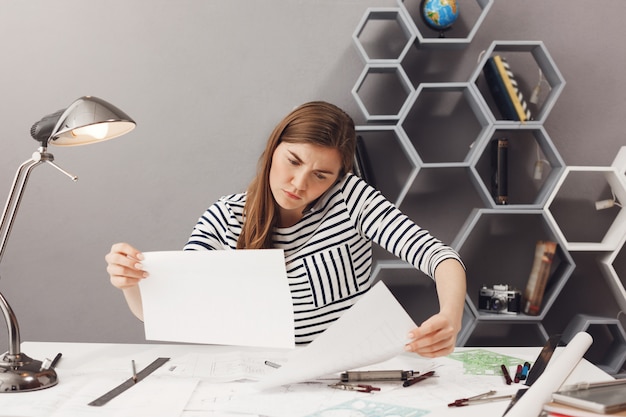 Biznes, niezależny, koncepcja pracy zespołowej. Młody przystojny zdezorientowany żeński architekt siedzi w coworking, rozmawia przez telefon z klientem i próbuje znaleźć informacje w dokumentach.
