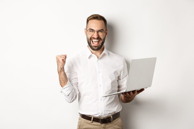 Biznes. Szczęśliwy menadżer wygrywający online, cieszący się pompką pięścią, trzymający laptopa i triumfujący, stojący