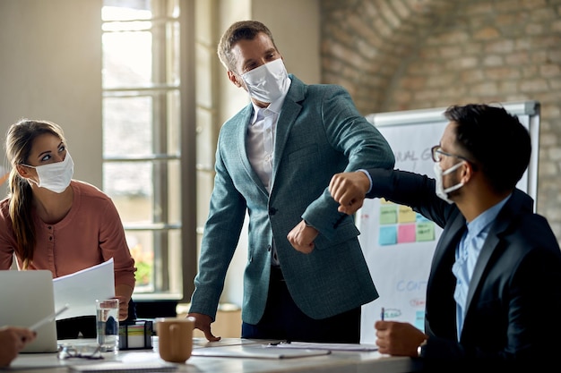 Biznesmen i jego współpracownik noszą ochronne maski na twarz i obijają się łokciem podczas powitania w biurze