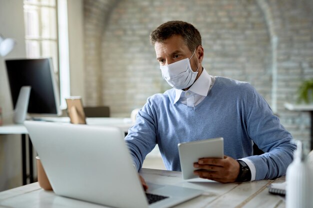 Biznesmen noszący maskę ochronną podczas pracy na komputerze i korzystania z cyfrowego tabletu w biurze