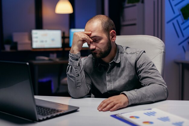 Biznesmen przepracowany śpi opierając głowę na dłoni. Pracoholik zasypiający z powodu pracy do późnych godzin nocnych sam w biurze przy ważnym dla firmy projekcie.