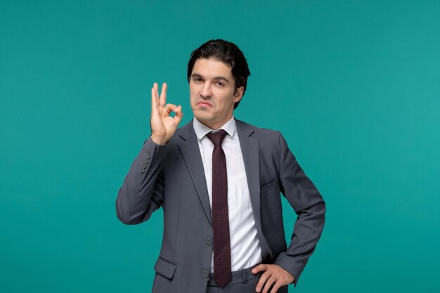 Biznesmen przystojny młody facet brunetka w szarym biurowym garniturze i krawacie pokazując znak ok