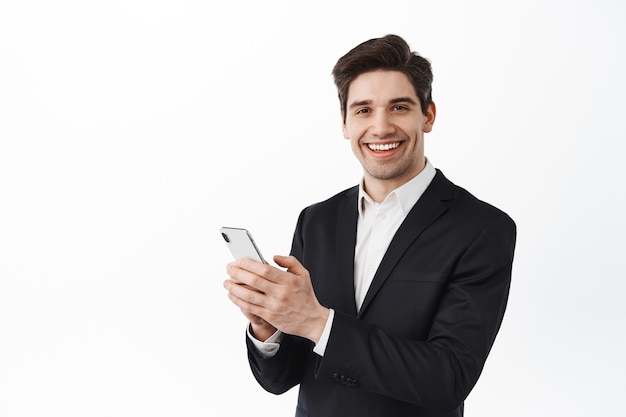 Biznesmen w czarnym garniturze korzystający z telefonu komórkowego, stojący ze smartfonem i patrzący z przodu, uśmiechnięta, biała ściana