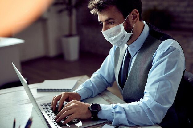 Biznesmen z maską na twarzy pracujący na laptopie w biurze podczas pandemii koronawirusa