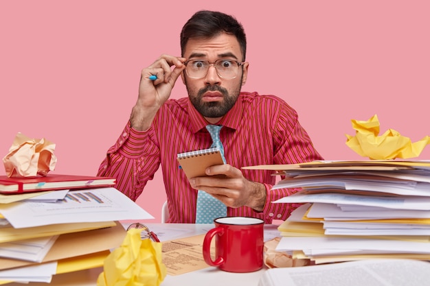 Bezpłatne zdjęcie biznesmen z przestraszonym wyrazem twarzy czuje się sfrustrowany, ma na biurku arkusze finansowe, trzyma notatnik, pije kawę, nosi formalną koszulę