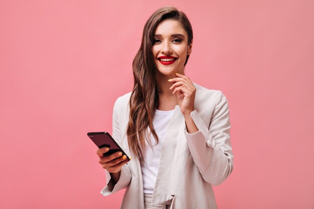 Biznesowa dama z czerwonymi ustami trzyma telefon na różowym tle. Kędzierzawa brunetka w stroju biurowym uśmiecha się i patrzy na kamerę.