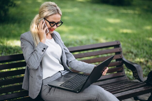 Biznesowa kobieta opowiada na telefonie w parku na ławce z laptopem
