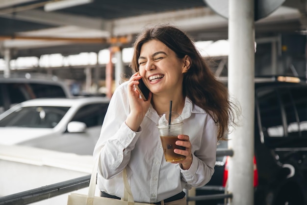 Biznesowa młoda kobieta z kawą rozmawia radośnie przez telefon na parkingu