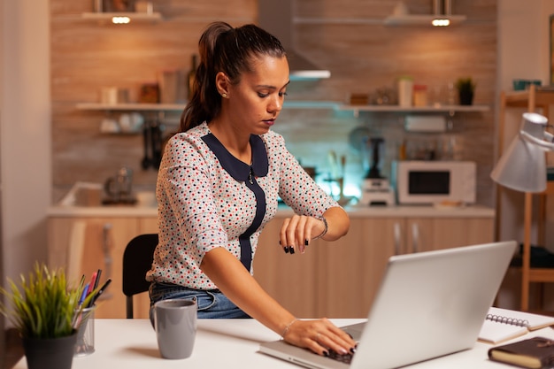 Bizneswoman sprawdza czas na zegarku podczas pracy z domu pracownika przy użyciu nowoczesnych technologii w połowie...