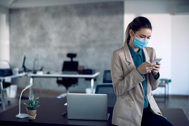 Bizneswoman z ochronną maską na twarz wysyła SMS-y na telefon komórkowy w biurze
