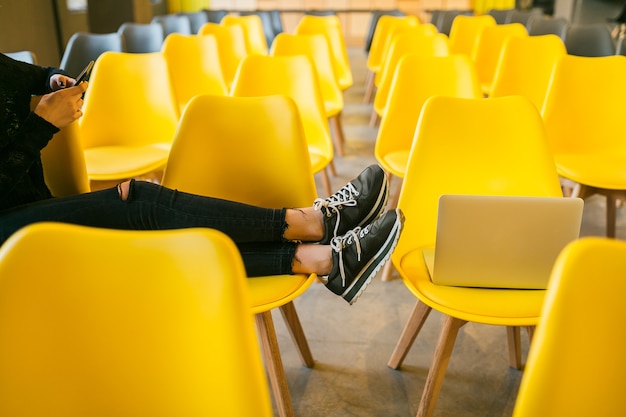 Bliska nogi młodej stylowej kobiety siedzącej w sali wykładowej z laptopem, sala lekcyjna z wieloma żółtymi krzesłami, trampki obuwie, trend w modzie buty