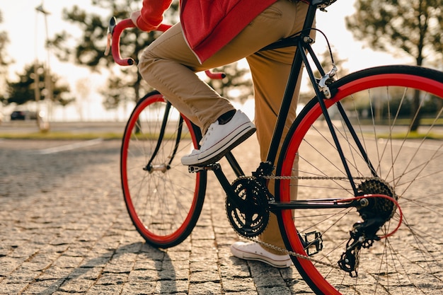 Bliska nogi w trampkach i ręce na kierownicy w stylu hipster brodaty mężczyzna w czerwonej bluzie z kapturem i beżowych spodniach jedzie samotnie z plecakiem na rowerze zdrowy aktywny styl życia podróżnik z plecakiem