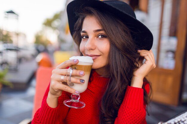 Bliska piękna młoda brunetka kobieta w stylowym czarnym kapeluszu i jasnoczerwonym swetrze siedzi w kawiarni na otwartej przestrzeni i pić kawę z mlekiem lub cappuccino w słoneczny poranek.