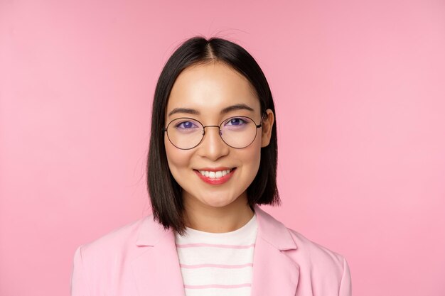 Bliska portret azjatyckiej kobiety korporacyjnej profesjonalnej bizneswoman w okularach uśmiechniętej i patrzącej pewnie na różowe tło aparatu