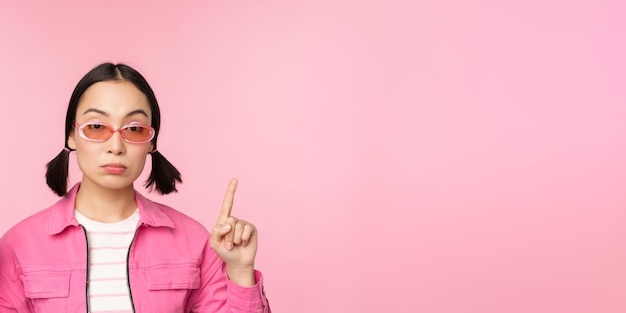 Bliska portret azjatyckiej stylowej dziewczyny patrzącej sceptycznie palcem wskazującym w górę z nierozbawioną twarzą stojącą na różowym tle