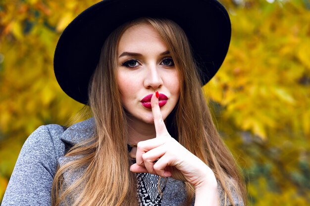 Bliska portret eleganckiej ładnej blondynki kobiety pozującej w jesienny zimny dzień w parku miejskim, ubrana w elegancki czarny kapelusz, długie włosy, jasny makijaż