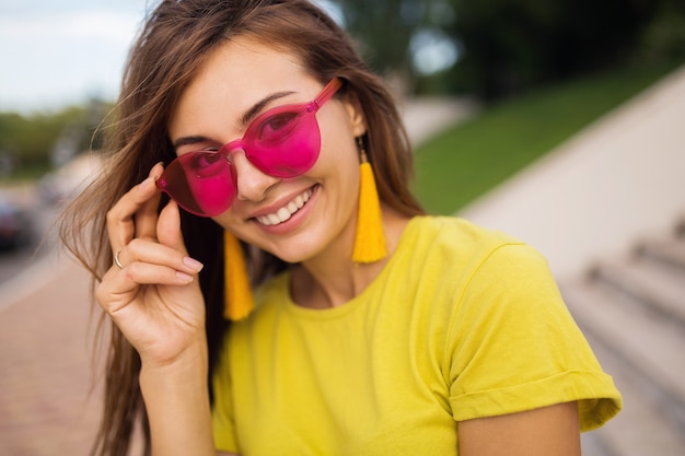 Bliska portret młodej atrakcyjnej uśmiechniętej kobiety bawiącej się w parku miejskim, pozytywna, szczęśliwa, ubrana w żółty top, kolczyki, różowe okulary przeciwsłoneczne, trend w modzie lato, stylowe akcesoria, kolorowe