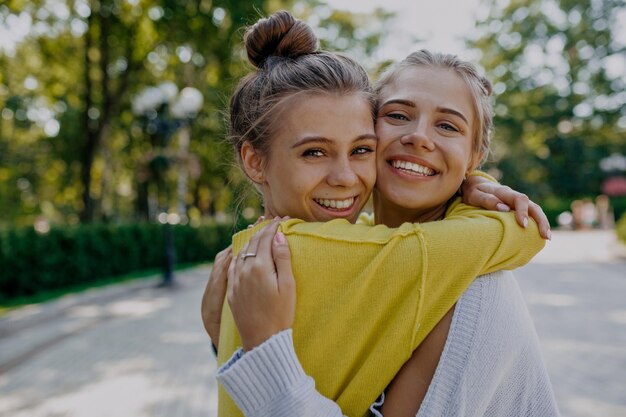 Bliska portret młodej uroczej damy z jasnobrązowym sianem i wspaniałym uśmiechem ma na sobie żółty sweter przytulający siostrę w niebieskim swetrze na tle zielonego słonecznego parku
