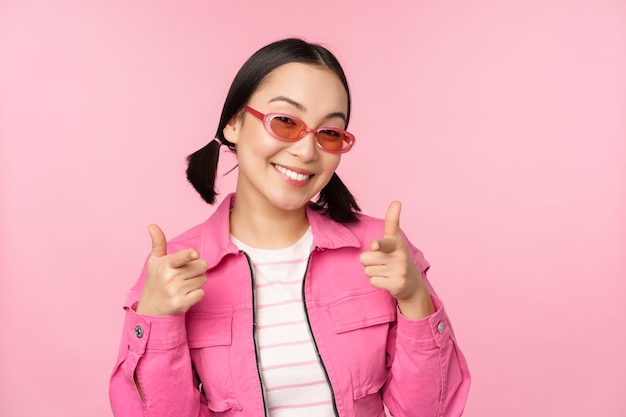 Bliska Portret Nowoczesnej Azjatyckiej Dziewczyny W Okularach Przeciwsłonecznych Uśmiechający Się Wskazującymi Palcami Na Aparat Chwali Cię, Zapraszając Lub Komplementując Stojąc Na Różowym Tle