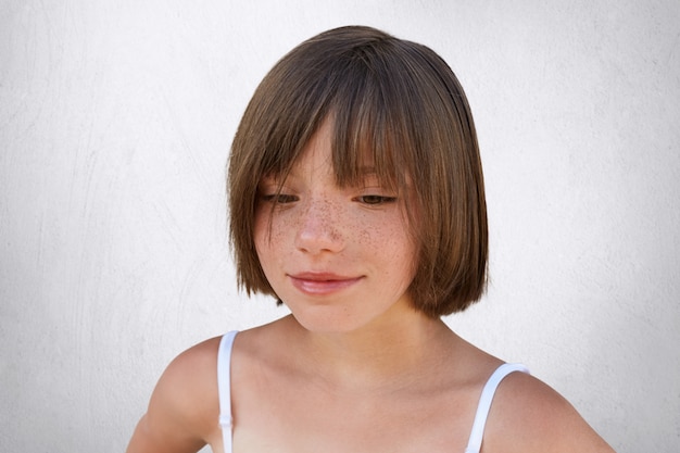 Bliska portret piegowatego dziecka z krótką stylową fryzurą, patrzącego w dół i marzącego o czymś przyjemnym. Piękna dziewczyna z konkretnym wyglądem pozuje nad biel ścianą