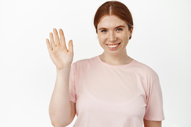 Bezpłatne zdjęcie bliska portret przyjaznej imbirowej dziewczyny ze szczerym uśmiechem, machająca ręką do kamery, przywitaj się, pozdrawiając klientów, witaj, wykonaj gest cześć fala, stojąc na białym tle