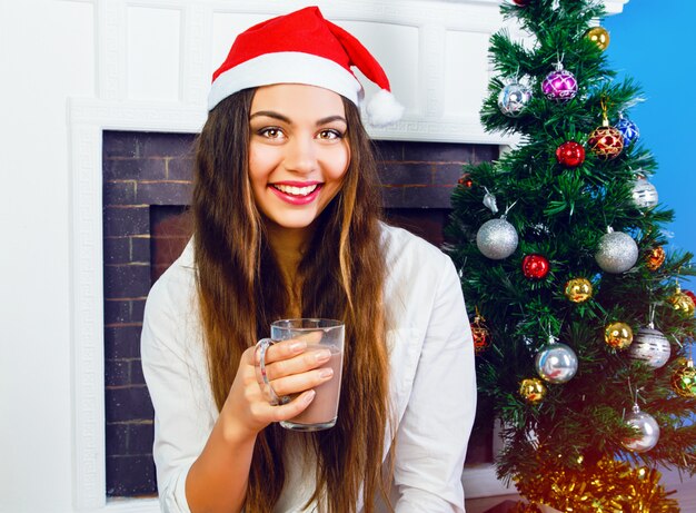 Bliska portret styl życia ładna brunetka kobieta pije gorącą czekoladę w sylwestra, ubrana w santa hat i siedząc przy kominku i ozdobione choinką. Przytulna domowa atmosfera.